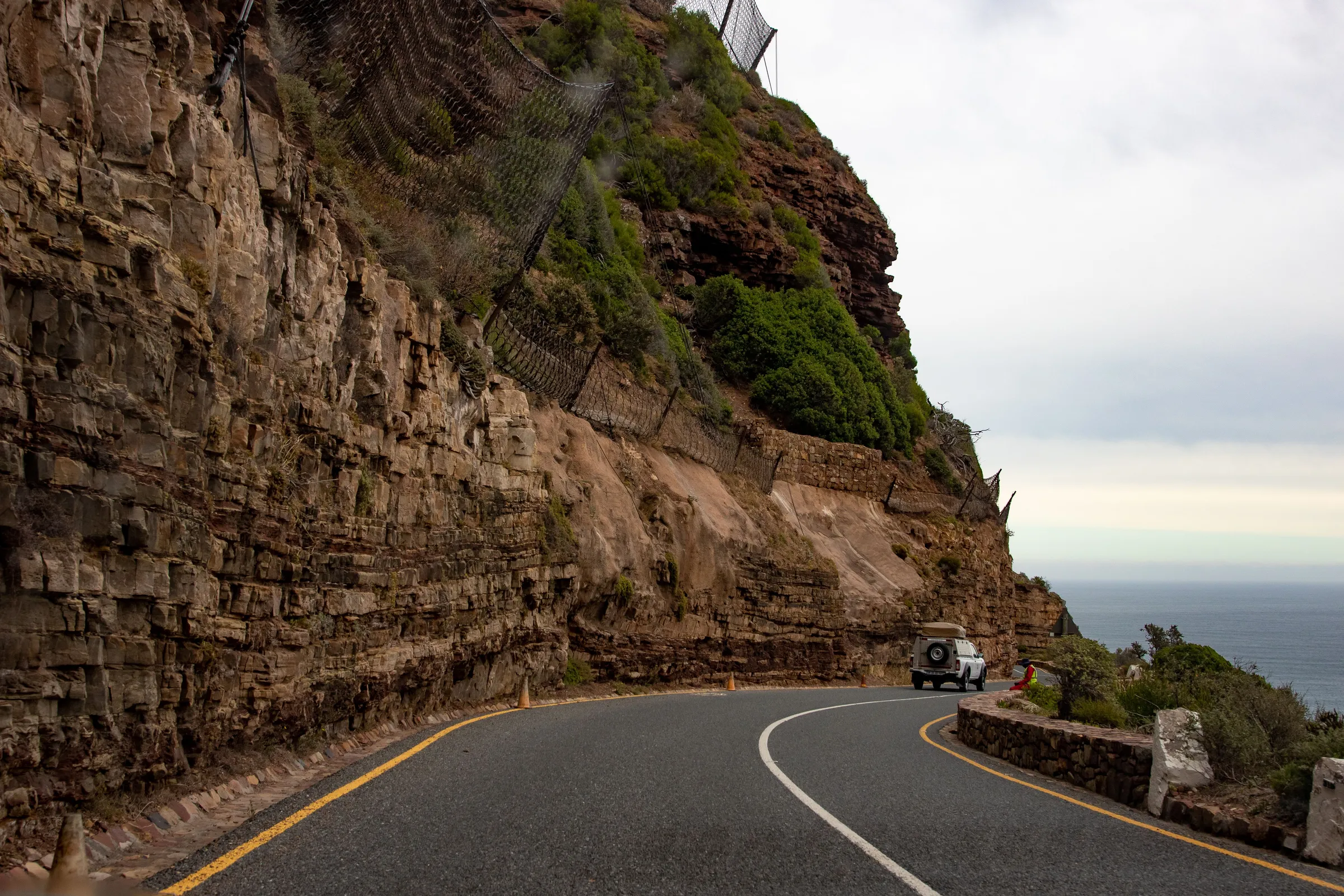 A pickup on a curving road cut below layered cliffs hung with rockfall netting, with the sea to the right