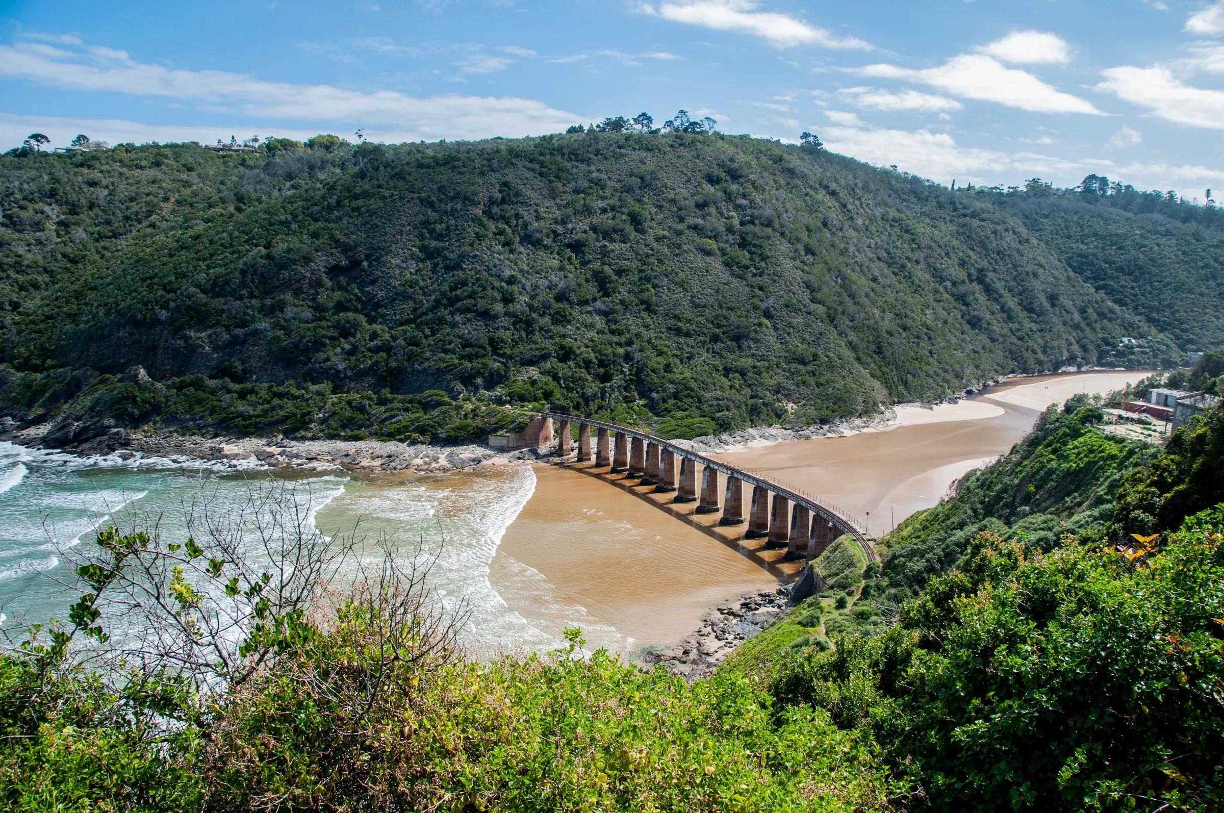 A river mouth with a railway bridge and a beach below forested hills