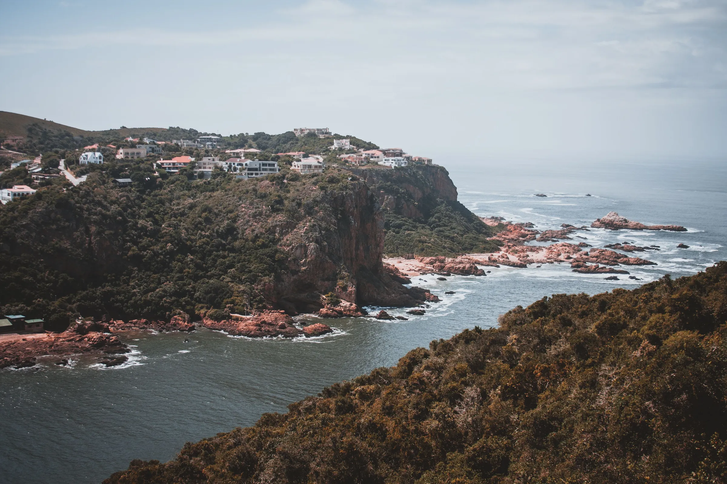 A headland with houses above rocks and a calm sea inlet