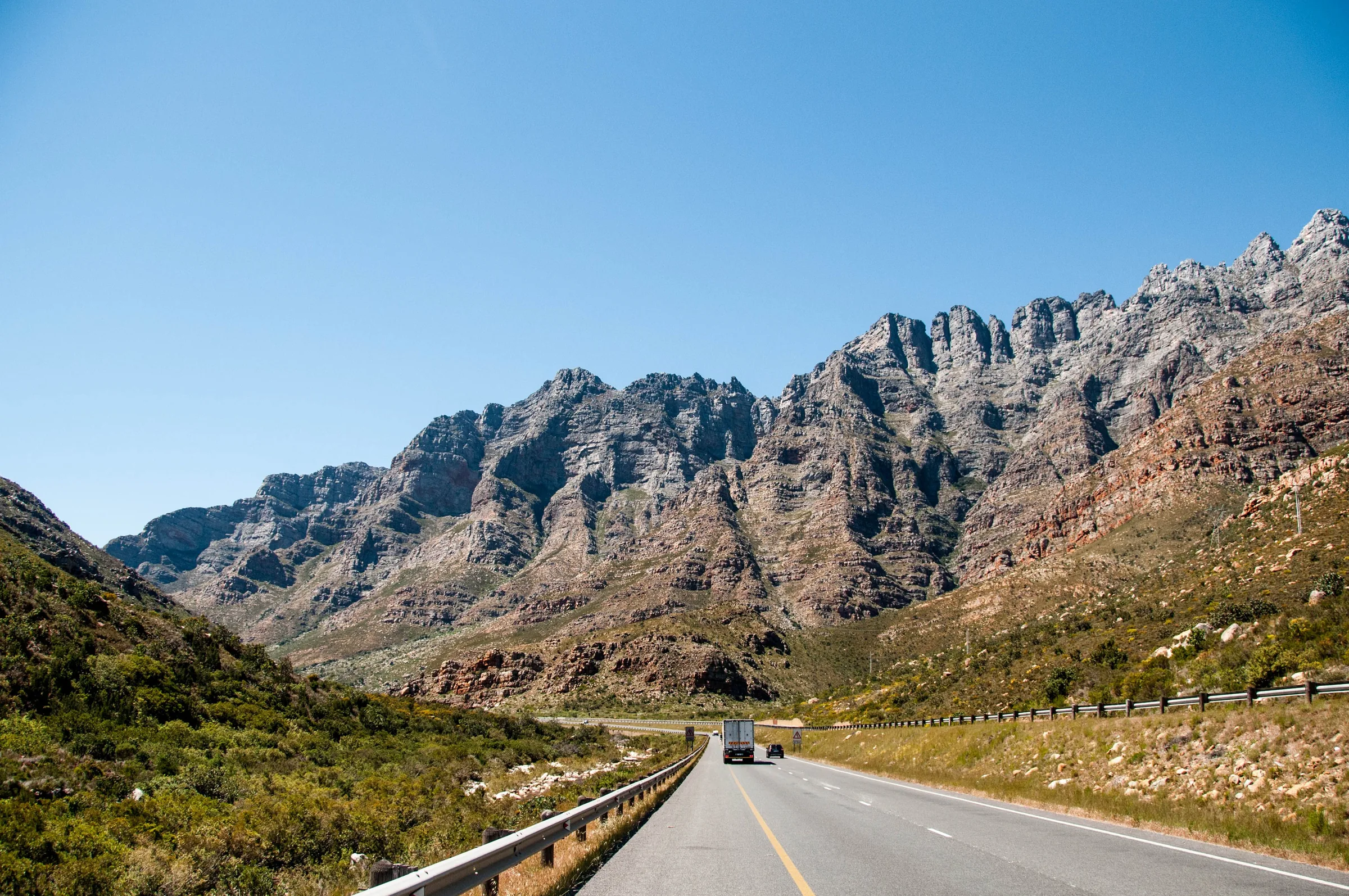 A truck and a car keeping left on a road below jagged rocky mountains